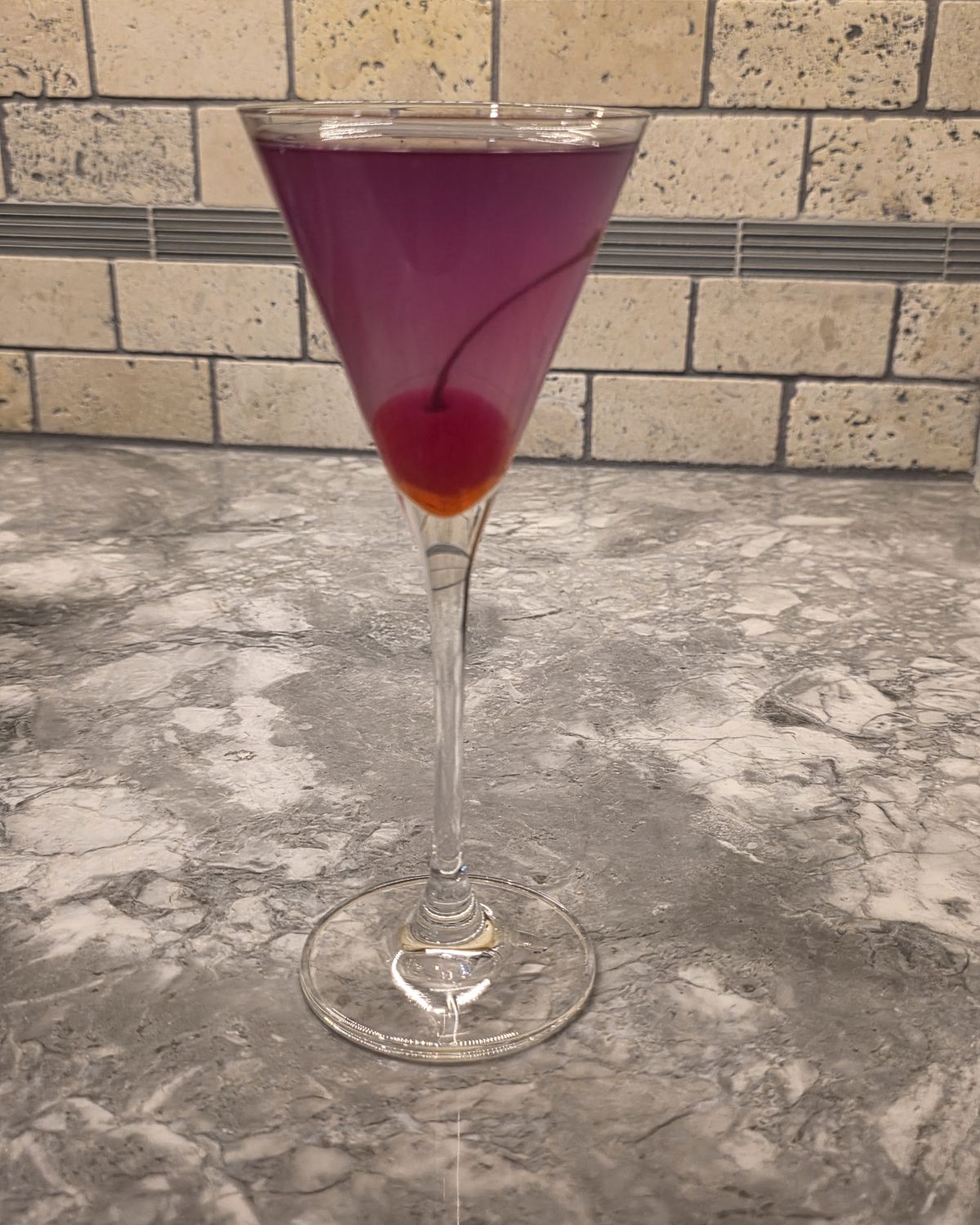Tall stemmed martini glass holding the finished Aviation cocktail with maraschino cherry, photographed against the bar's travertine backsplash