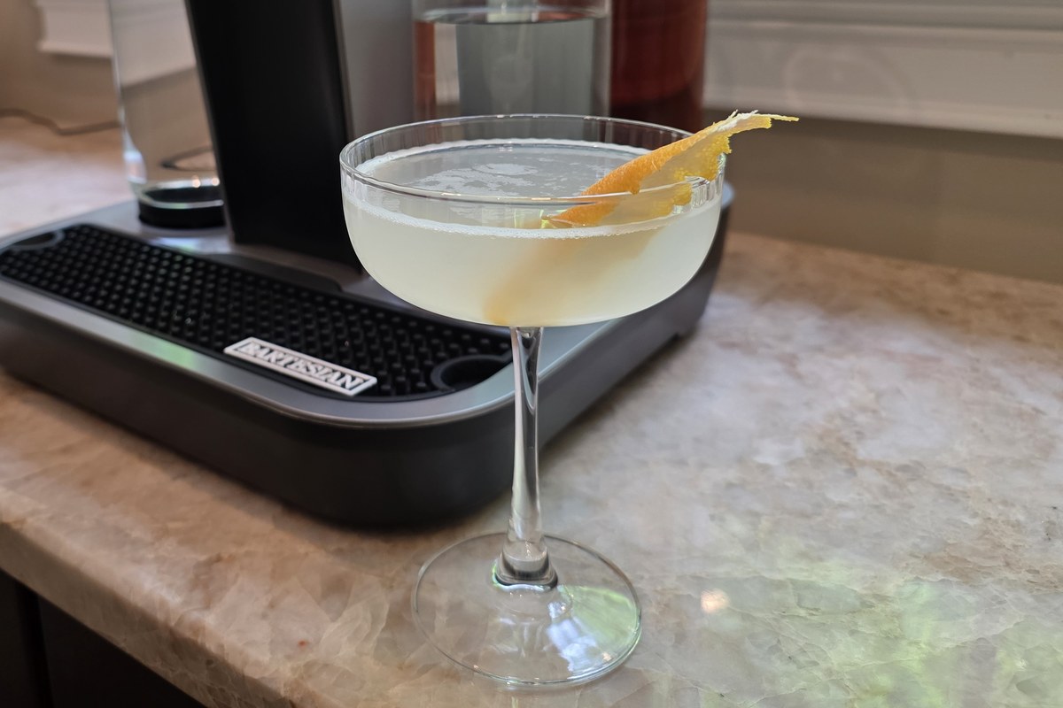 The finished Bee's Knees cocktail in a coupe glass with a lemon peel, beside the Bartesian machine on a home bar