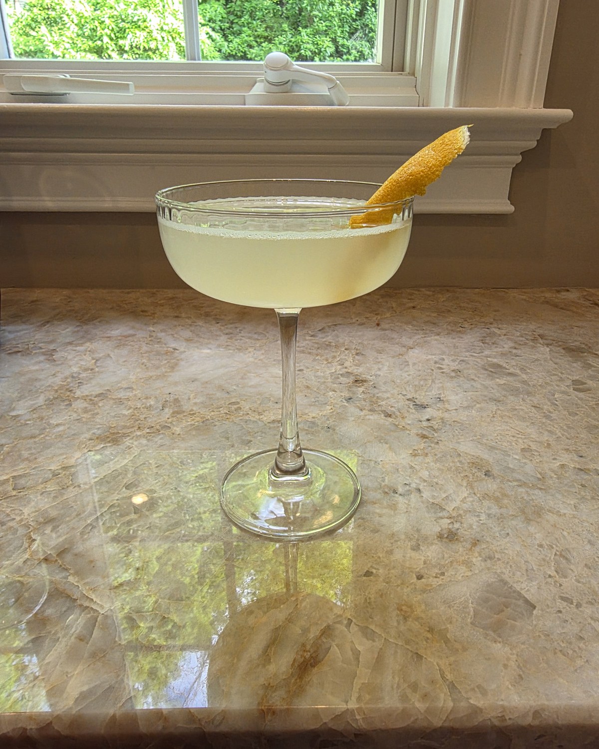 Bartesian Bee's Knees cocktail in a coupe glass with a pale yellow color and a lemon garnish on a home bar