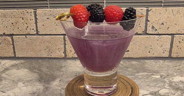 A purple, shimmering Witch's Heart cocktail in a stemless martini glass with raspberries and blackberries on a pick across the rim