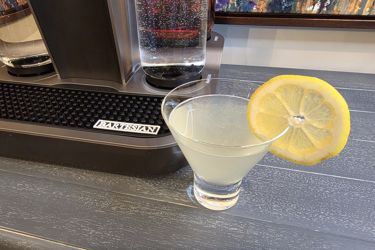 The finished Lemon Drop cocktail in a stemless martini glass with a lemon wheel, beside the Bartesian machine on a home bar