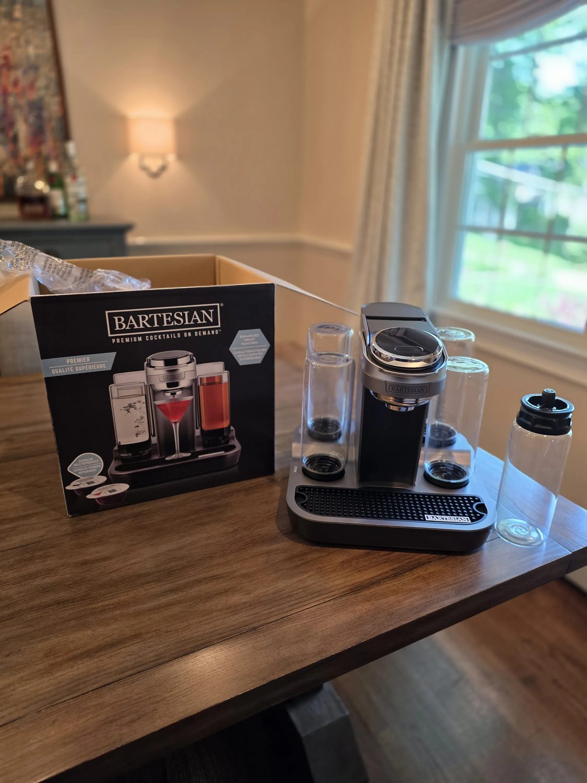 Bartesian Premier cocktail machine beside its retail box on a wood table, with glass spirit bottles in place and a tumbler