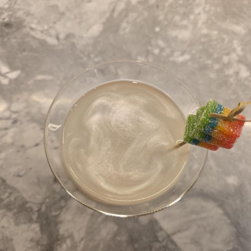 Looking down into a glass of Sour Spell, with a swirl of shimmer in the pale drink and rainbow sour candy on a pick at the rim