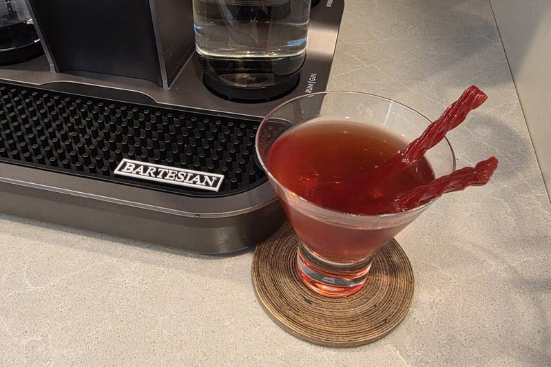 A Vampire's Kiss in a stemless martini glass with two red licorice twists, on a wooden coaster beside a Bartesian Premier