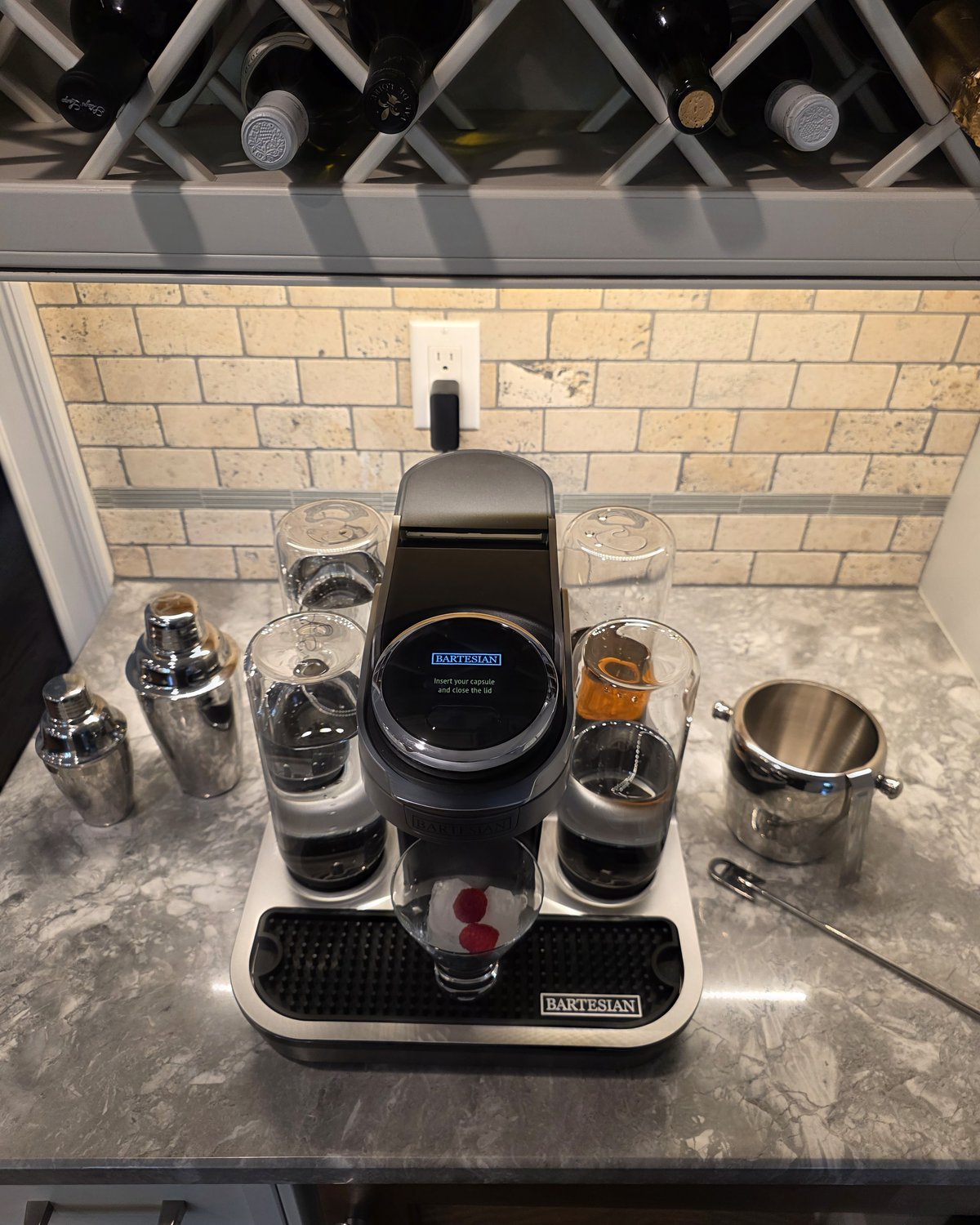 Bartesian Professional cocktail machine ready for first use, with stainless cocktail shaker and ice bucket alongside, glass with raspberry garnish in place under the spout, travertine backsplash and wine rack visible above