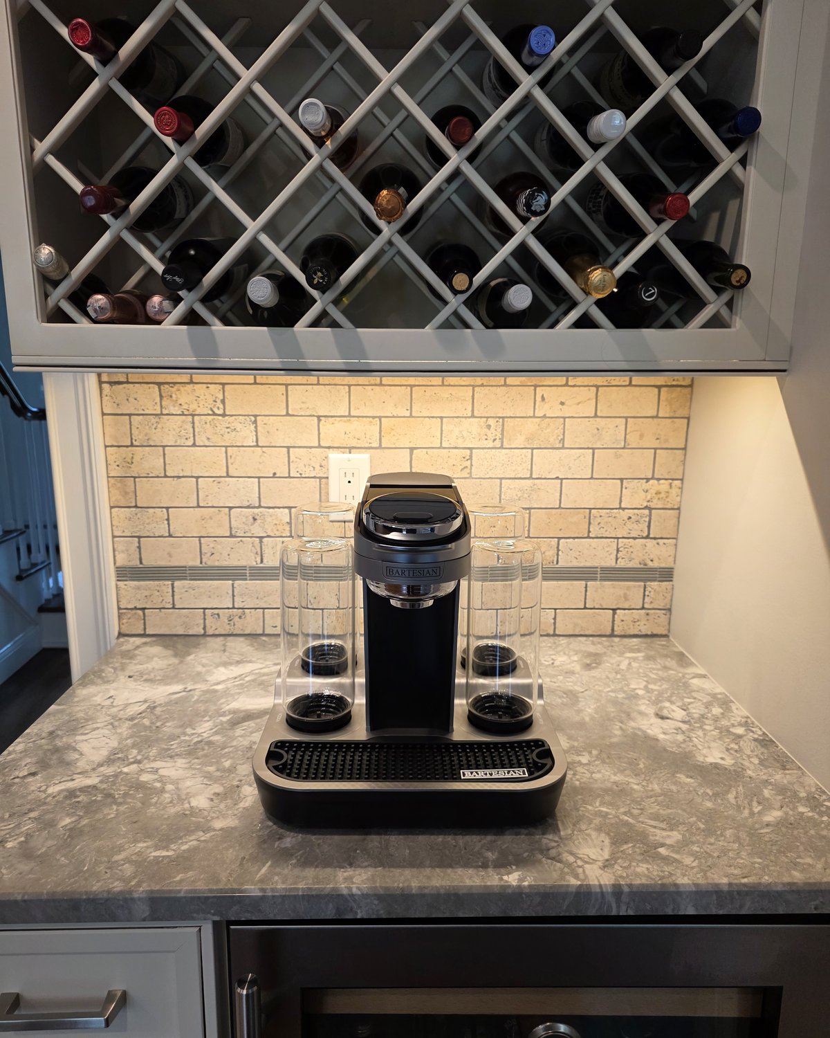 Bartesian Professional machine fully unboxed and placed on its final bar location, empty spirit bottles upside down in slots, travertine subway-tile backsplash, wine rack visible above