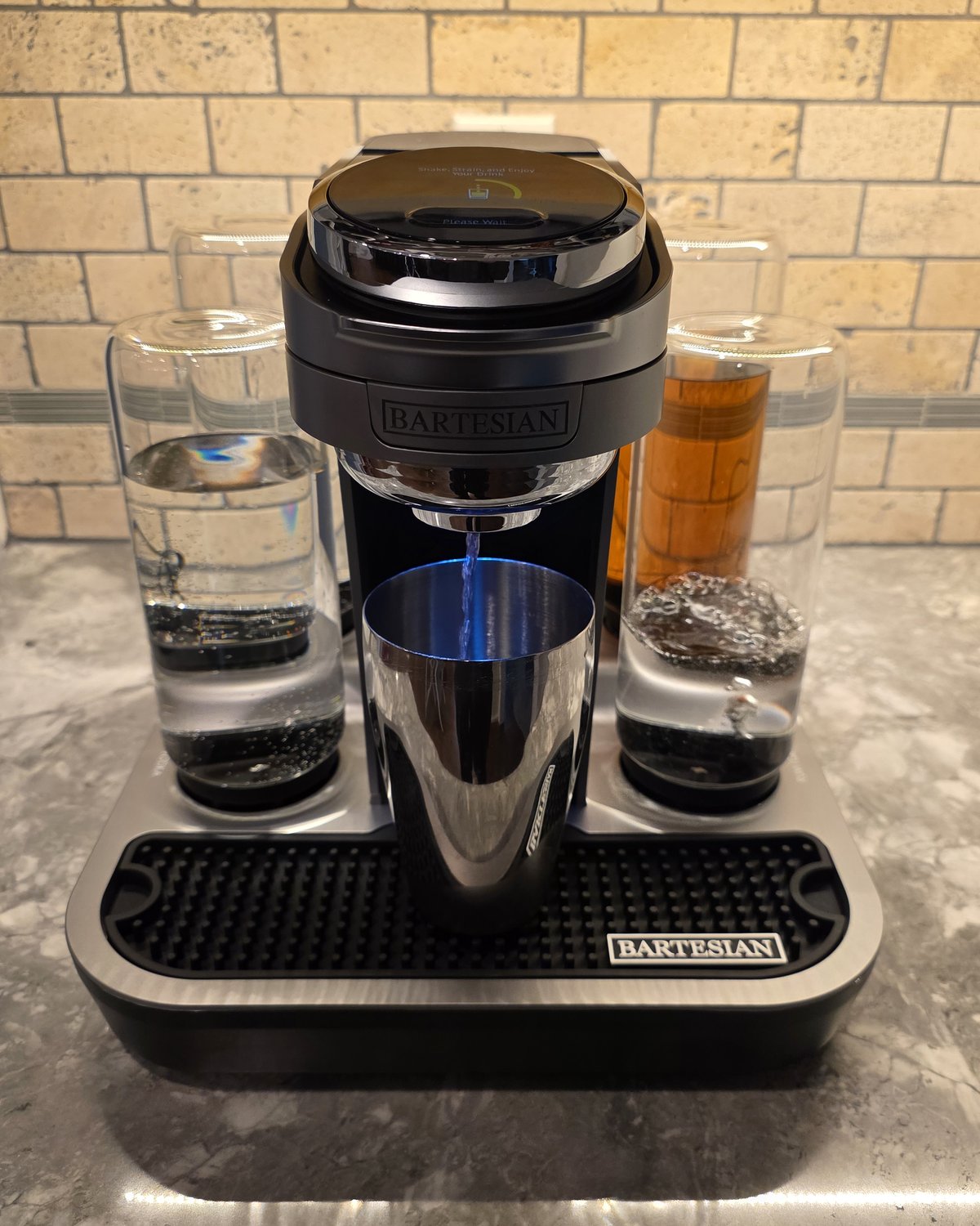 Bartesian Professional actively dispensing a cocktail, blue liquid pouring from the spout into a stainless steel cocktail shaker, screen displaying Shake Strain and Enjoy