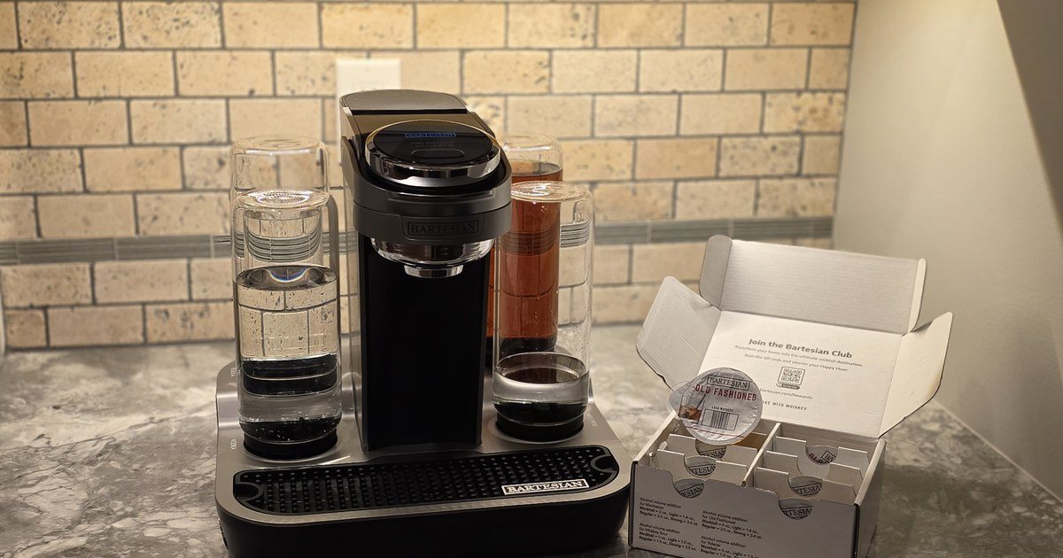 Bartesian cocktail machine on a granite countertop next to an open Bartesian Whiskey Lovers Collection variety pack, with an Old Fashioned pod visible inside the box ..... stone subway tile backsplash and warm under-cabinet lighting in the background.