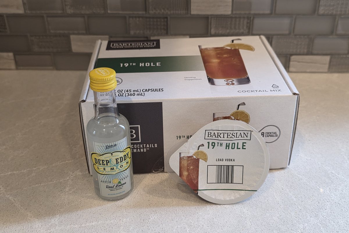 A Deep Eddy Lemon flavored vodka mini bottle in front of a Bartesian 19th Hole pod box on a kitchen counter