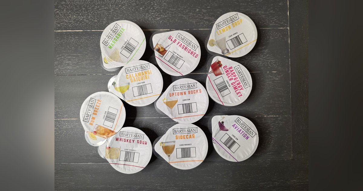 Twelve Bartesian cocktail pods arranged on a dark wood table, each capsule printed with its cocktail name and a LOAD VODKA, LOAD GIN, LOAD WHISKEY, LOAD RUM, or LOAD TEQUILA label showing the spirit is added separately.