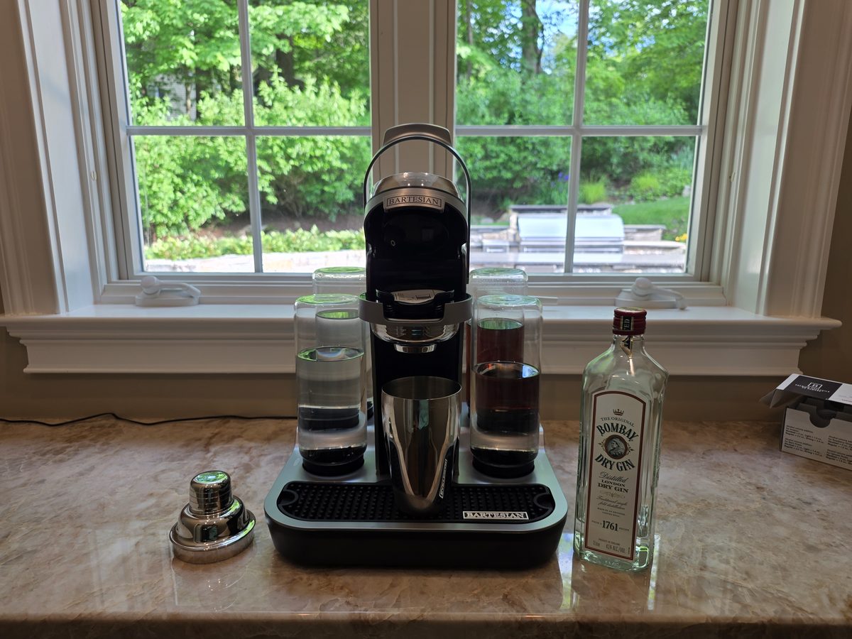 A Bartesian cocktail machine on a marble kitchen counter with its four glass spirit bottles in the holder, a steel shaker, and a bottle of Bombay London Dry Gin beside it, in front of a bright garden window.