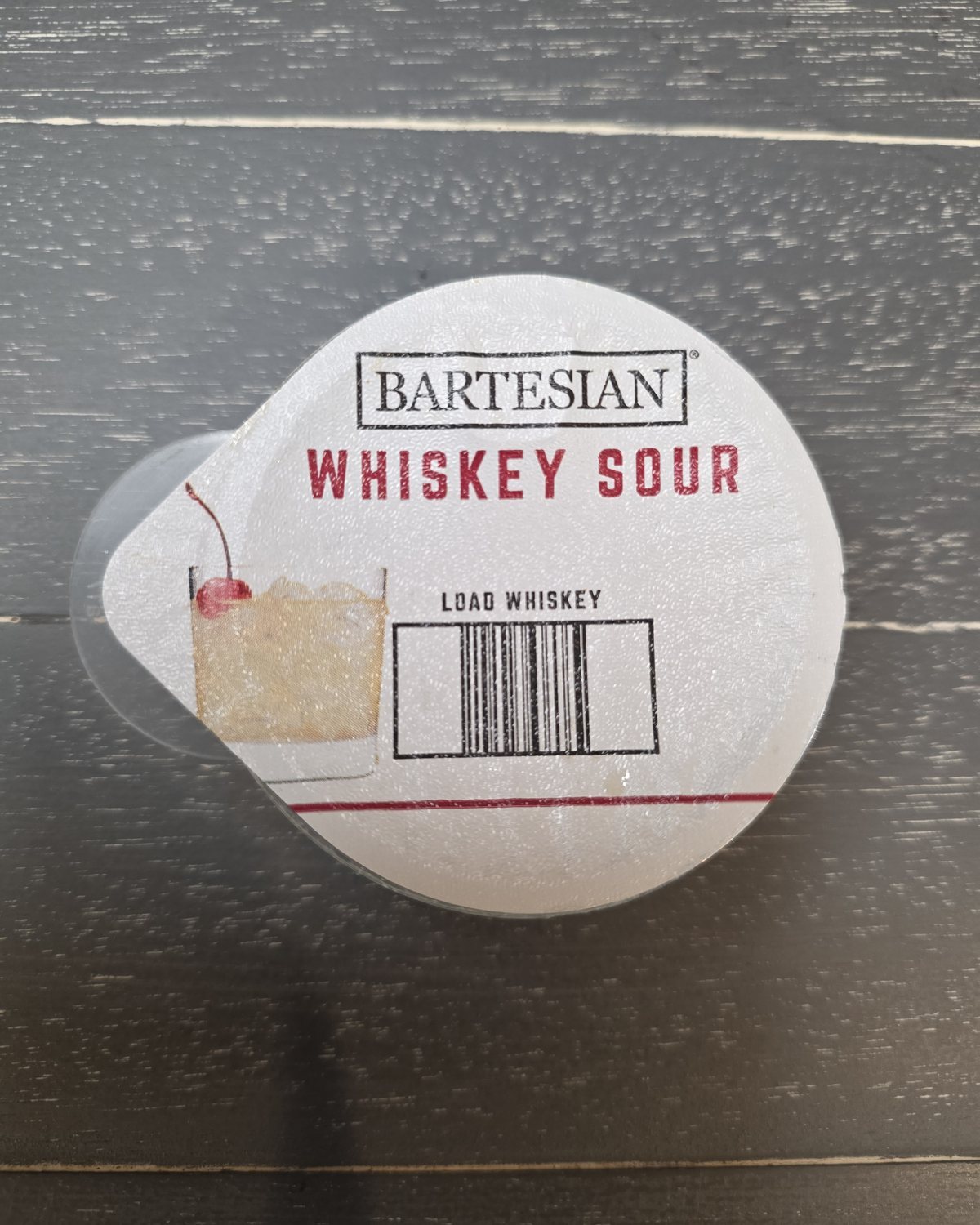 Top-down close-up of a Bartesian Whiskey Sour pod showing the printed barcode and LOAD WHISKEY label the machine scans to identify the cocktail