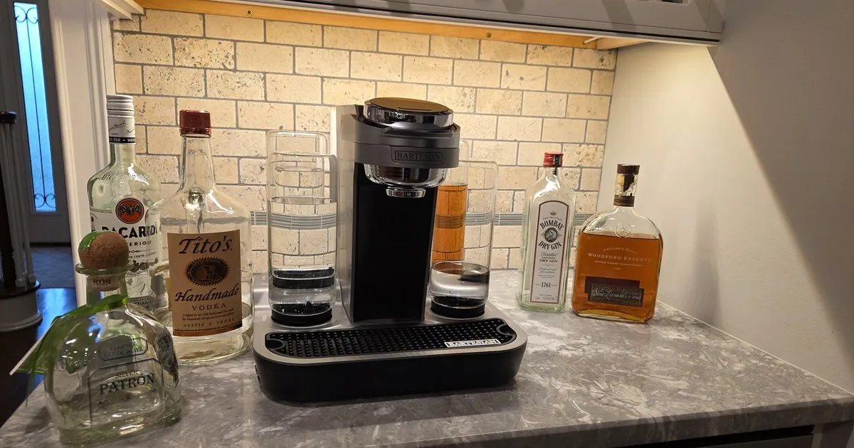 Bartesian cocktail machine on a granite countertop surrounded by Patrón, Bacardi, Tito's, Bombay Dry Gin, and Woodford Reserve ..... a hosting-ready home bar setup with stone backsplash and warm under-cabinet lighting.
