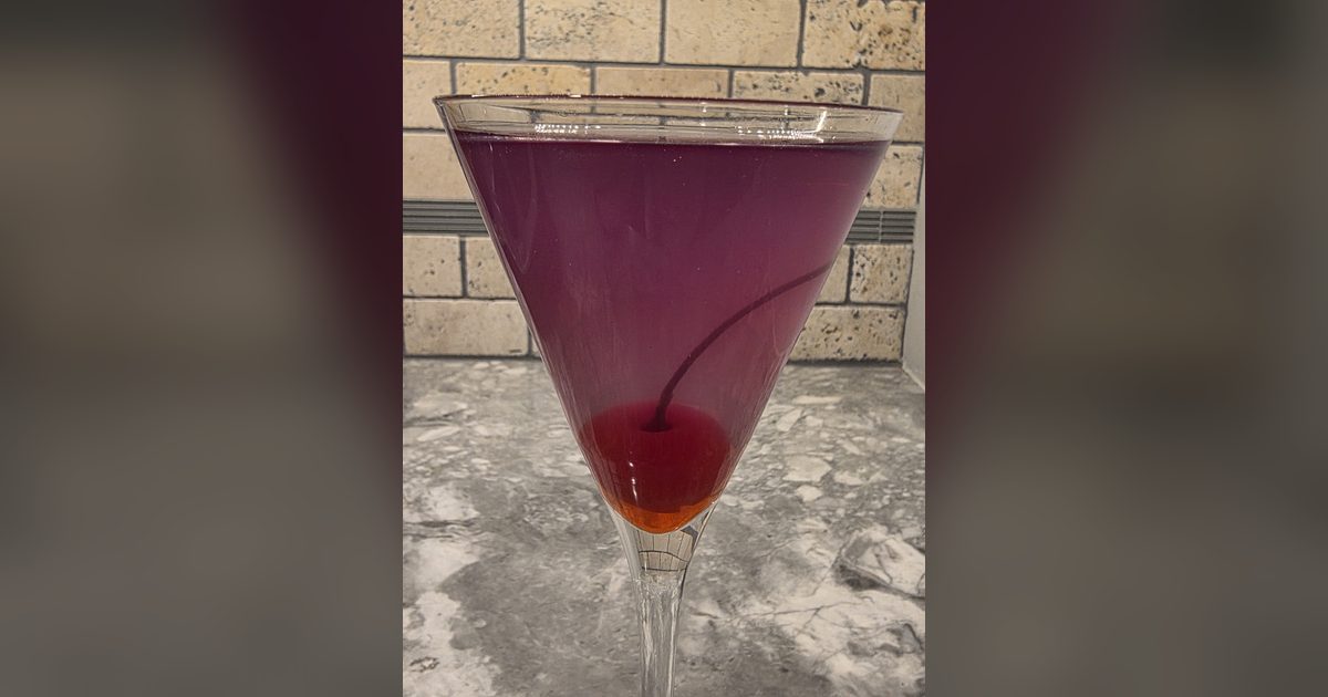 A deep purple Aviation gin cocktail with a maraschino cherry in a tall martini glass on a marble counter.
