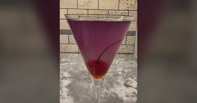 A deep purple Aviation gin cocktail with a maraschino cherry in a tall martini glass