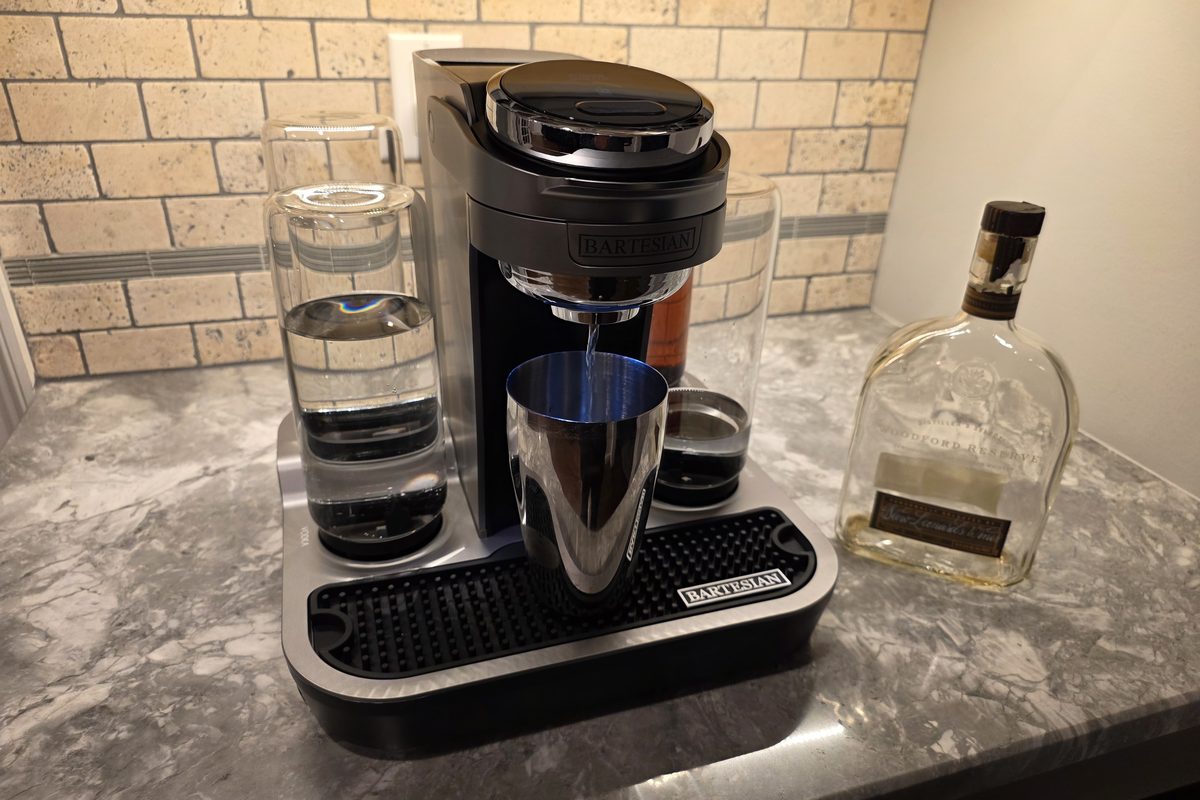 A Bartesian cocktail machine pouring a Manhattan into a steel shaker tin, with a bottle of Woodford Reserve bourbon beside it