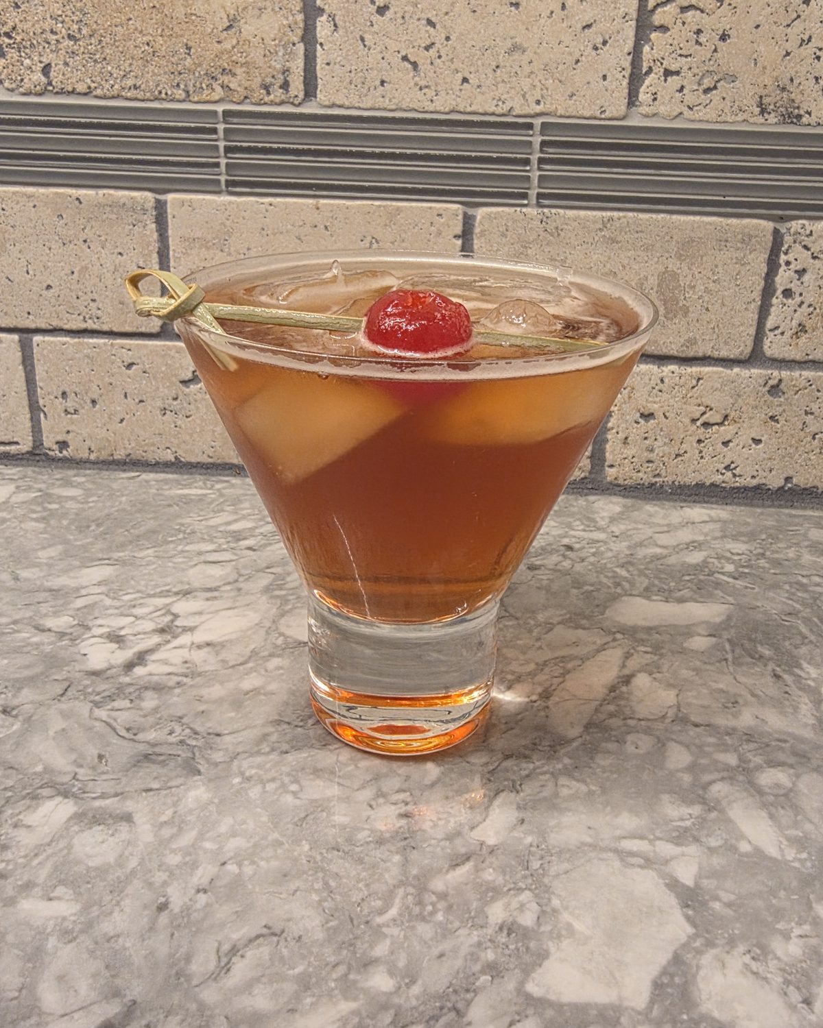 Bartesian Manhattan cocktail in a stemless martini glass over ice, garnished with a maraschino cherry, on a marble counter