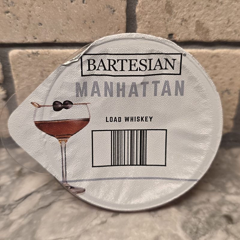 A Bartesian Manhattan cocktail capsule lid reading Load Whiskey, on a marble counter