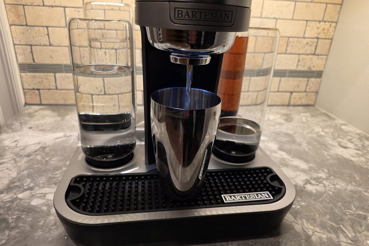 A Bartesian cocktail machine pouring an Old Fashioned into a steel cup, with a bottle of Woodford Reserve bourbon beside it