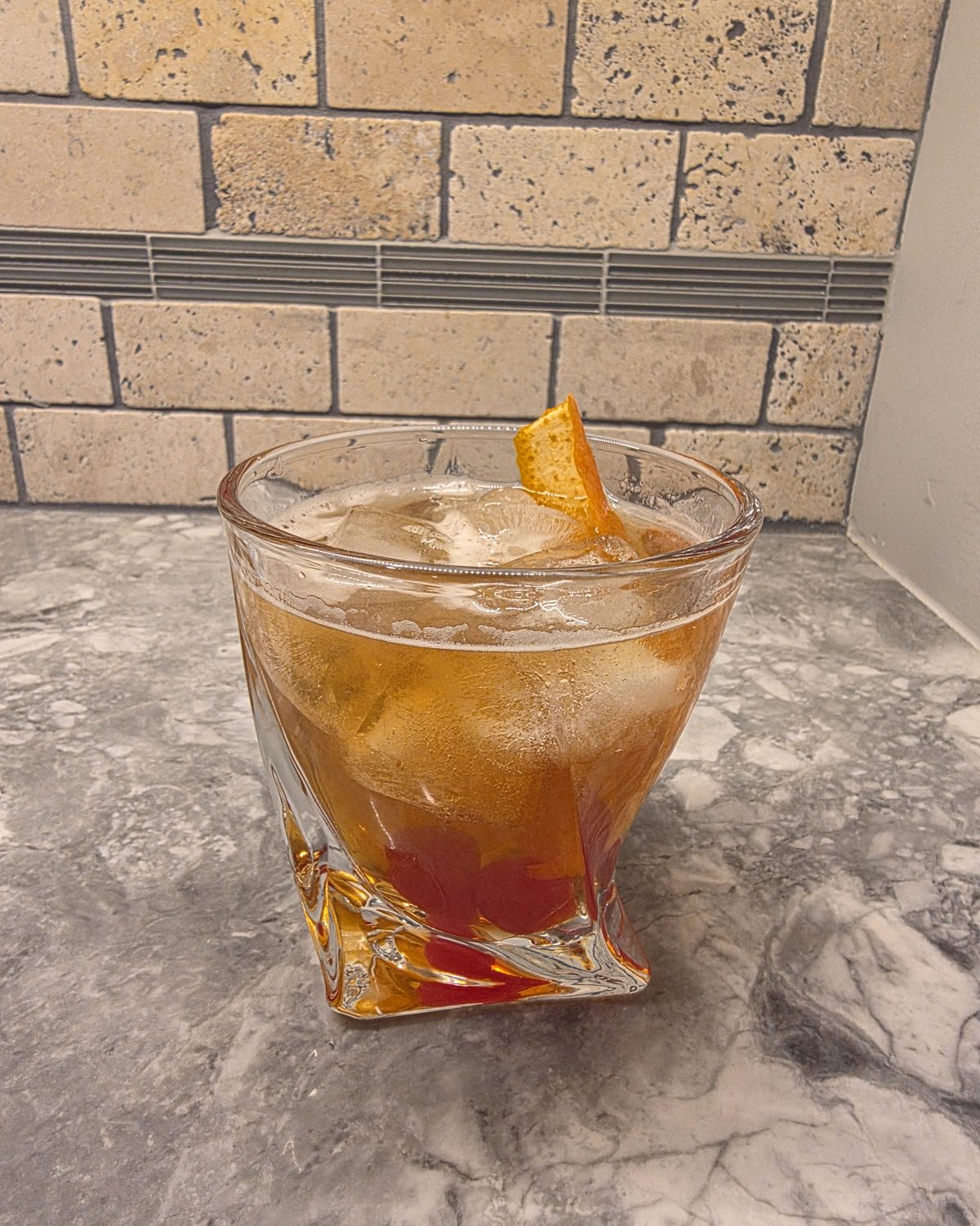 Bartesian Old Fashioned cocktail in a heavy rocks glass over ice, garnished with an orange peel and maraschino cherries on a marble counter