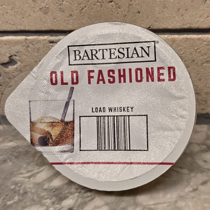 A Bartesian Old Fashioned cocktail capsule lid reading Load Whiskey, on a marble counter
