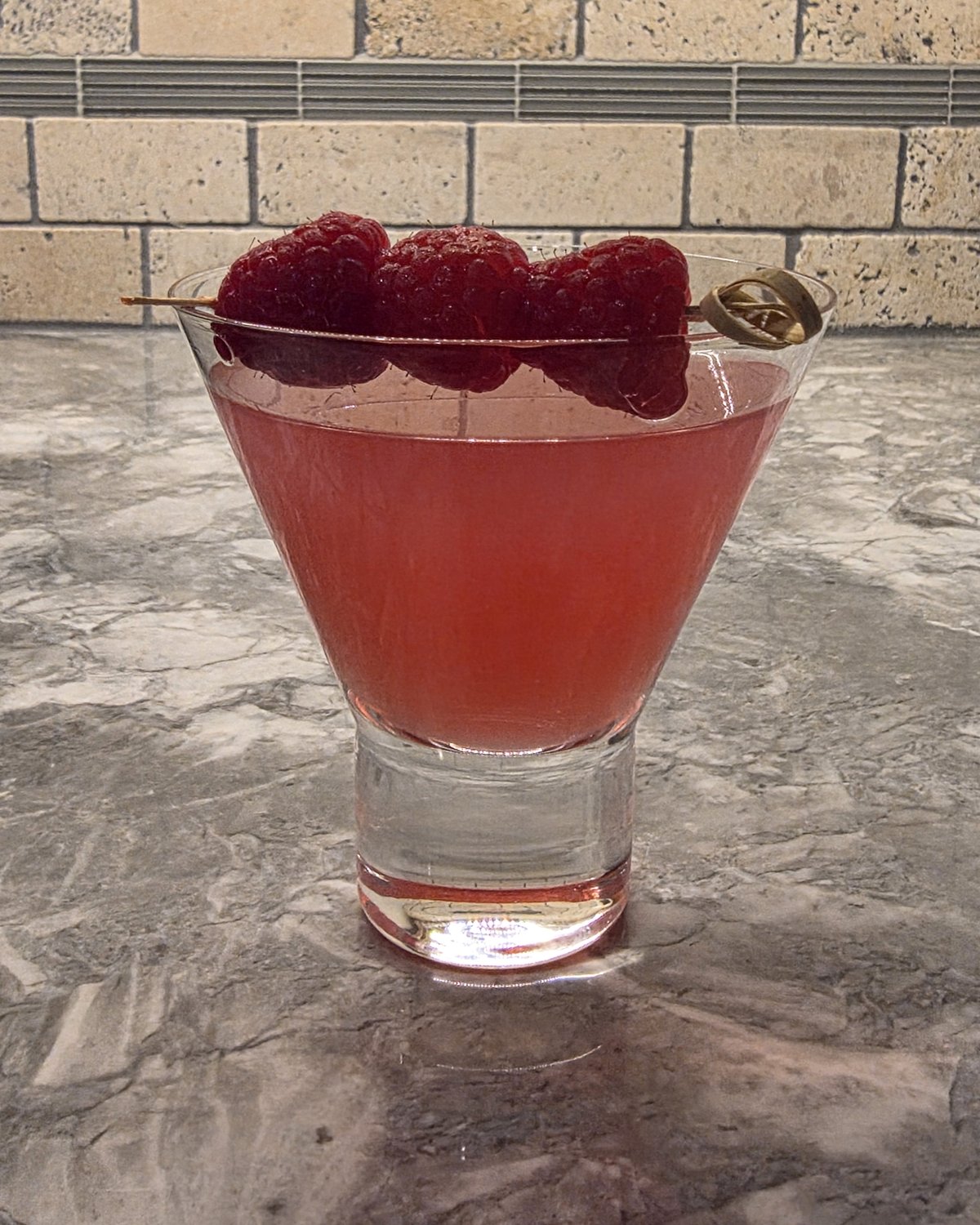 Close-up of the Raspberry Ginger Gimlet cocktail showing the bright pinkish-red color with three raspberries on a skewer garnish