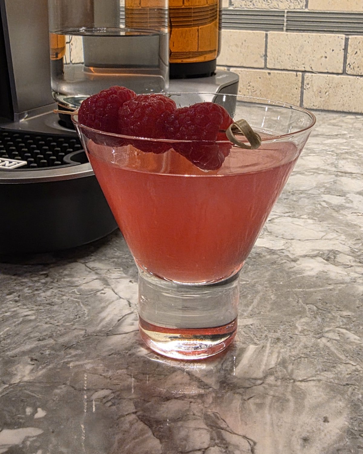 Raspberry Ginger Gimlet cocktail in a stemless martini glass on a marble counter with the Bartesian Pro machine softly out of focus in the background