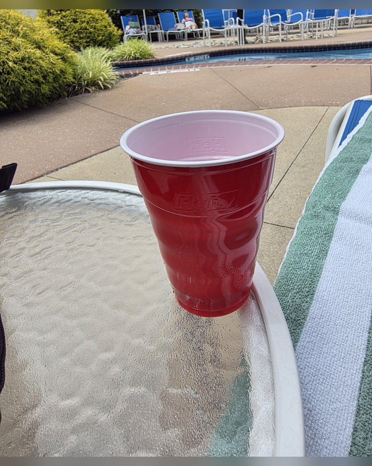 A red Solo cup with a cocktail on a glass poolside table at a resort, with lounge chairs, blue umbrellas, and the pool behind it