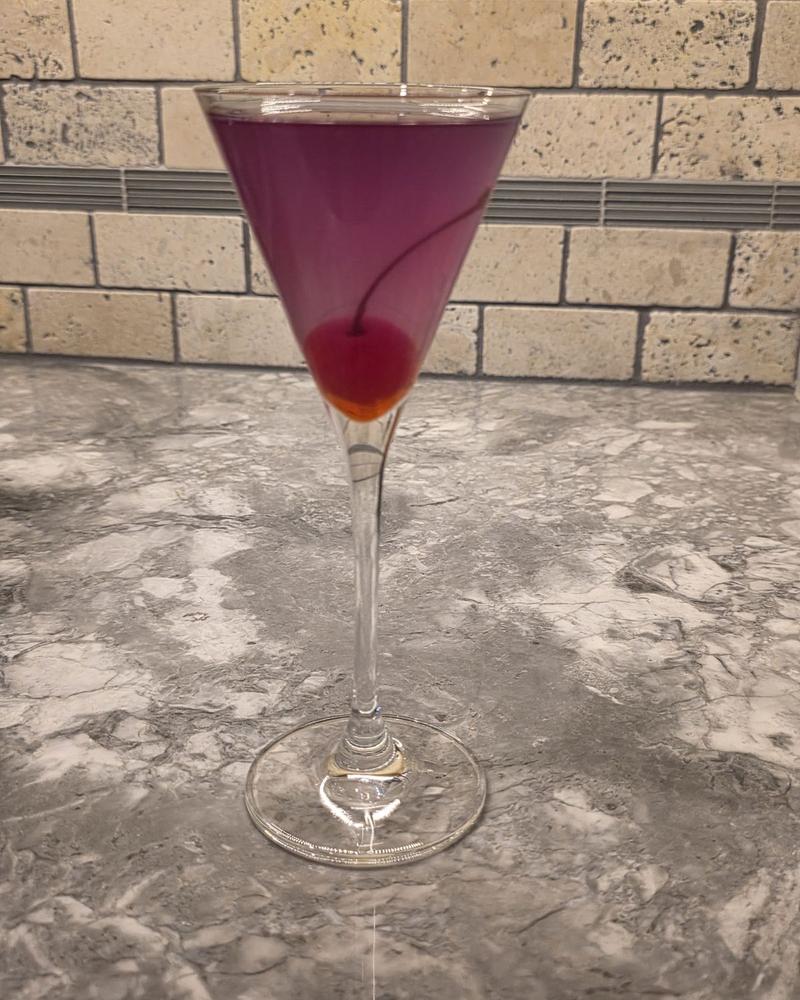 A deep purple Aviation with a cherry in a tall martini glass on a marble counter