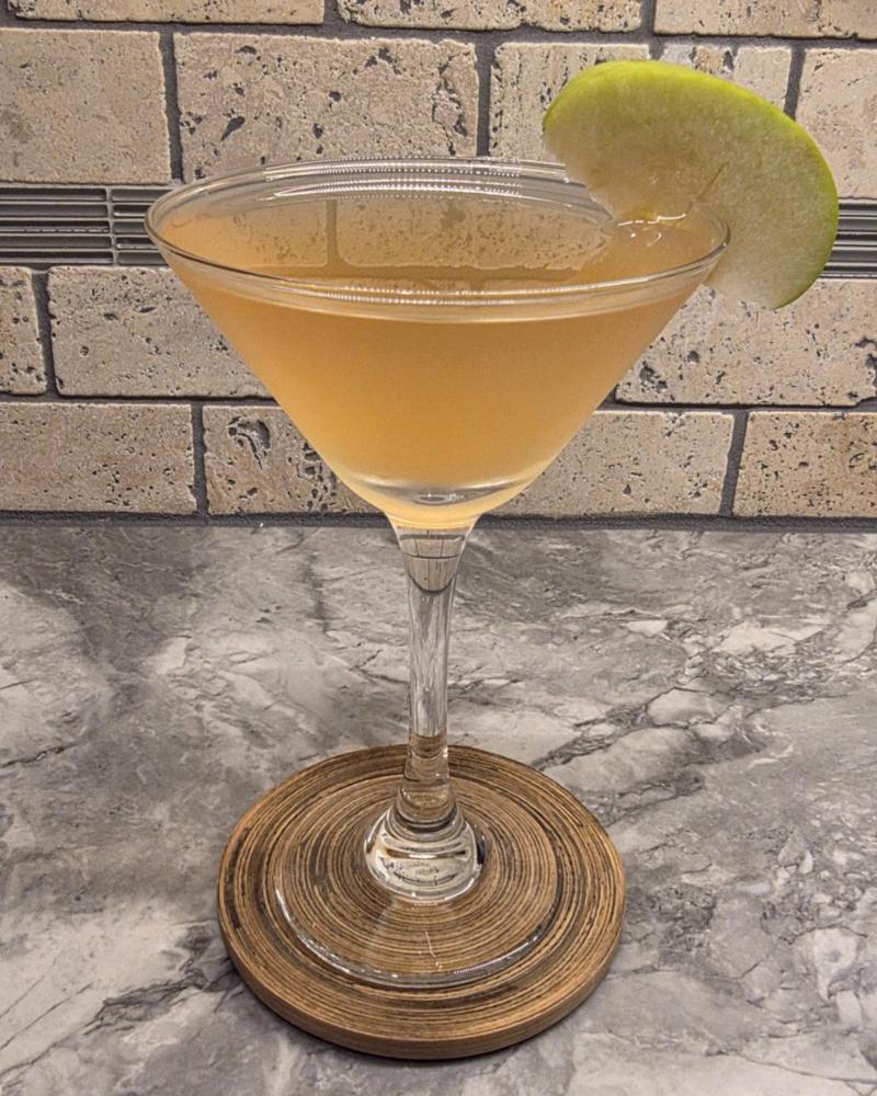 A golden Caramel Appletini in a martini glass with a green apple slice, on a wooden coaster