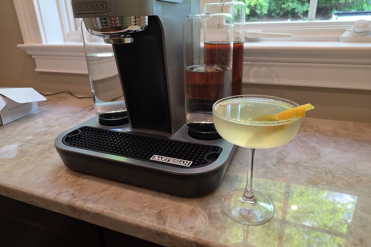 Finished Uptown Rocks cocktail in a coupe glass with a lemon peel, alongside the Bartesian machine by a sunlit window