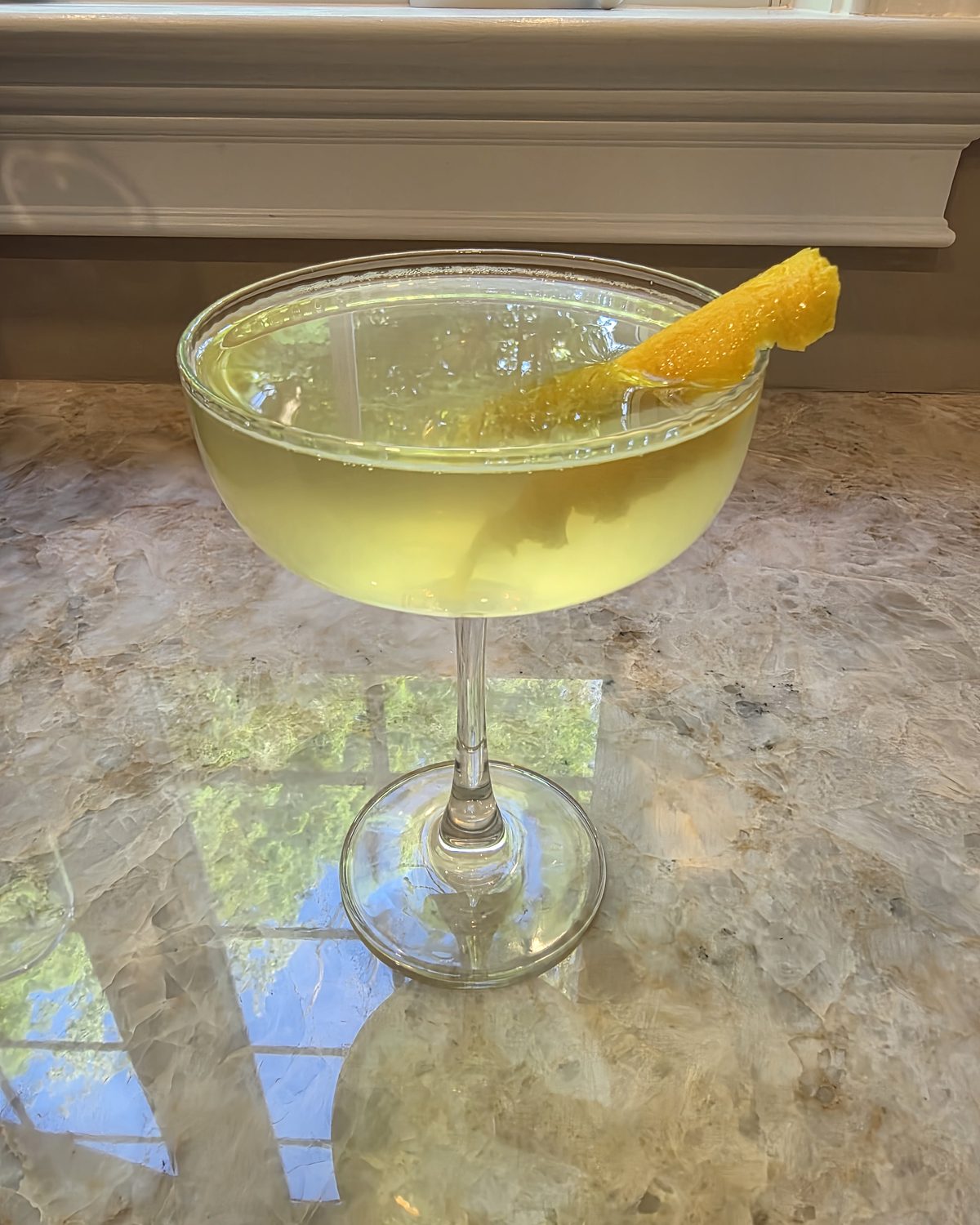 Bartesian Uptown Rocks cocktail in a coupe glass with a soft pale-yellow color and a lemon peel garnish on a marble counter by a sunlit window