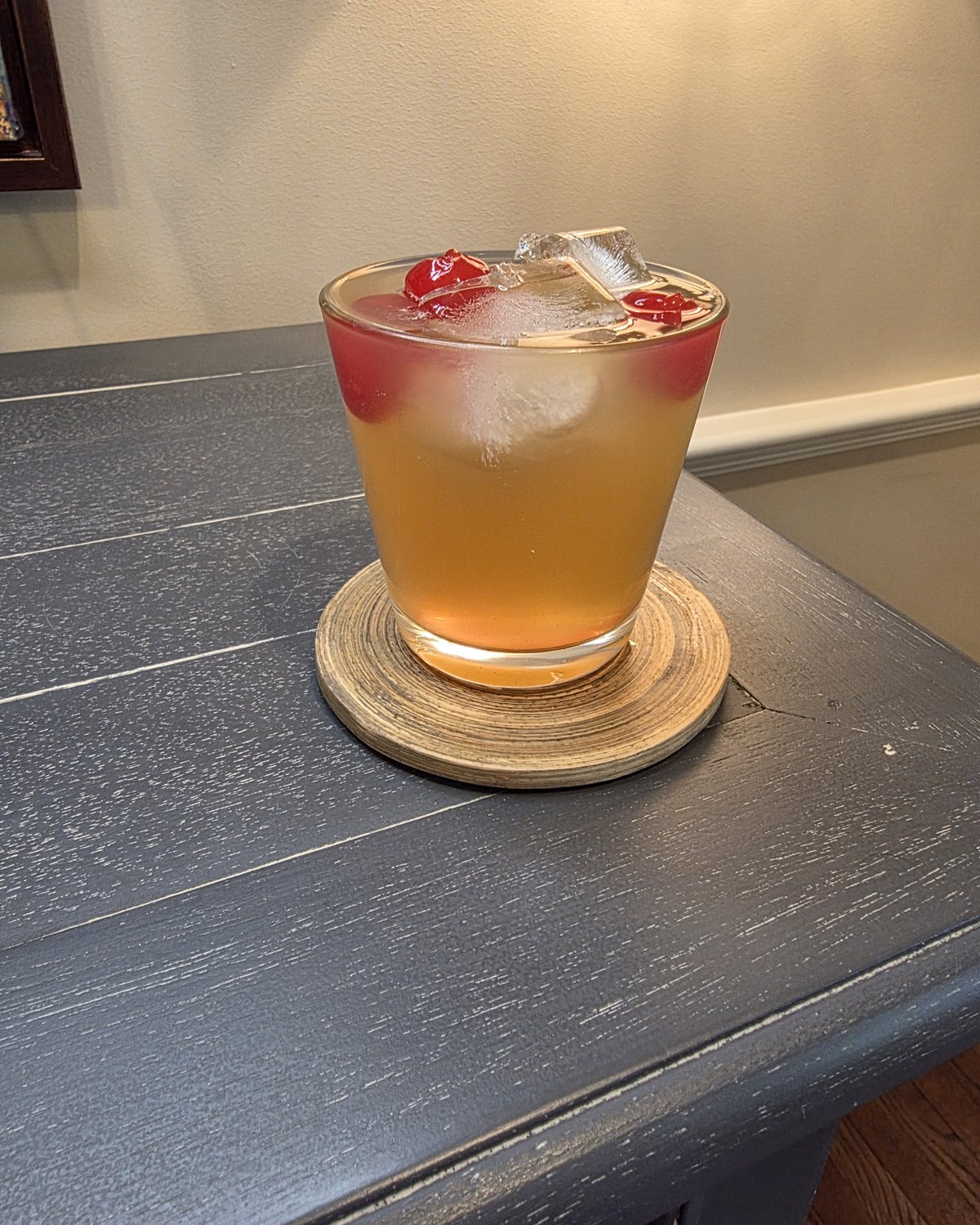 Bartesian Whiskey Sour cocktail in a low ball glass with maraschino cherries and ice on a wood coaster in a home bar corner