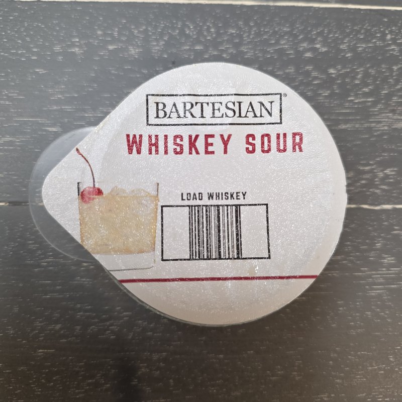 Bartesian Whiskey Sour capsule lid showing the cocktail illustration and the Load Whiskey instruction on a dark wood surface